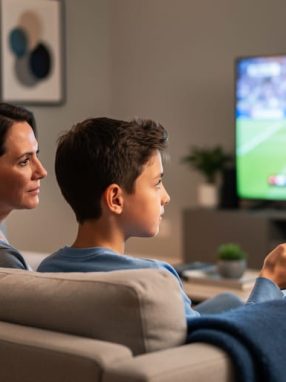 Parent holding a remote turns to speak with a preteen on a couch while a blurred sports game plays on the TV in the background under warm evening light.