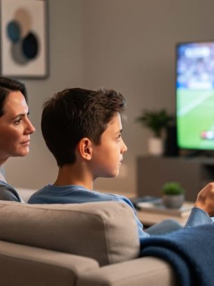 Parent holding a remote turns to speak with a preteen on a couch while a blurred sports game plays on the TV in the background under warm evening light.